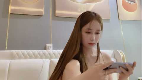 Media: Video of a young East Asian woman with long, straight brown hair, wearing a black top, sitting on a white couch, engrossed in a smartphone. Background features modern, abstract artwork and a neutral-colored wall.