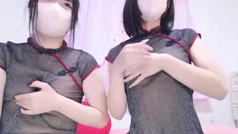 Media: Video of two young Asian women with fair skin, wearing sheer black cheongsams with red trim, covering their nipples with hands, in a bedroom with white walls and pink curtains.