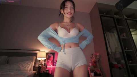 Media: Video of a confident Asian woman with medium build, light skin, and black hair, wearing a light blue off-shoulder top and white shorts, standing in a dimly-lit bedroom with a bed, shelves, and a lamp.