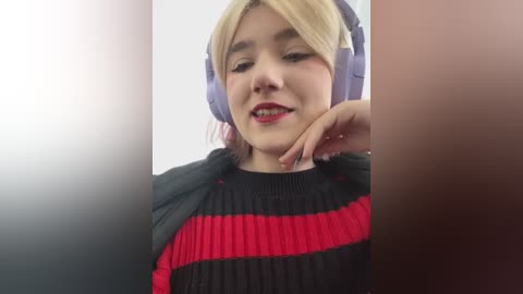 Media: Video of a young woman with light skin and blonde hair, wearing a black and red striped sweater, large purple headphones, and a slight smile, set against a blurred background.