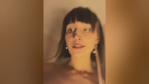 Media: Video of a topless young woman with fair skin and straight dark hair, wearing only silver hoop earrings, against a plain beige background. She has a neutral expression.