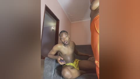 Media: Video of a shirtless Black man with a lean physique, wearing yellow shorts, playing a video game on a couch in a simple, peach-colored room with an orange curtain.