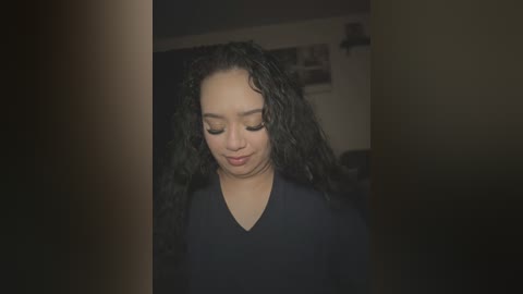 Media: A video of a young Black woman with long, curly black hair and light brown skin, wearing a black V-neck shirt, looking down thoughtfully in a dimly lit room.