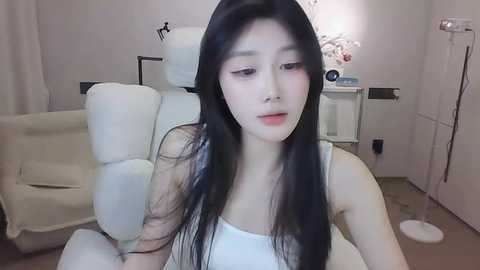 Media: Video of a young East Asian woman with long black hair, fair skin, and a slender build, wearing a white tank top, seated on a cream chair in a minimalist, softly lit room with a white wall and floral arrangement.