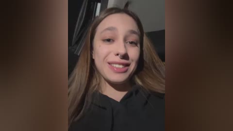 Media: Video of a young woman with fair skin, light brown hair, and a nose ring, smiling slightly. She wears a black top. Background is dimly lit, featuring a dark, indistinct room.