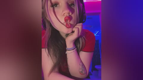 Media: Video of a young woman with long, dyed hair, wearing headphones, licking a red lollipop, in a dimly lit room with purple and pink lighting.