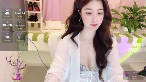 Media: Video of an East Asian woman with long, wavy black hair, wearing a white lace bra and robe, sitting in a bedroom with a green-striped bed, shelves, and plants.