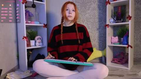 Media: Video of a red-haired, fair-skinned girl in a red-and-black striped sweater, white leggings, meditating on a turquoise mat in a whimsical, decorated room with shelves adorned with holiday ornaments and stuffed toys.