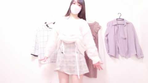 Media: Video of a young Asian woman with long black hair, wearing a white sweater and plaid skirt, standing in a room with a white wall and two hanging outfits.