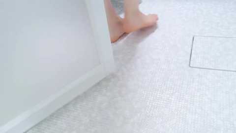 Media: Video of a corner in a room with white walls and light gray, textured flooring. A person\u2019s bare foot is partially visible, standing on the floor. The image has a soft, diffuse lighting.