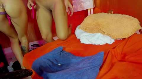 Media: Video of a woman with a light brown complexion, wearing a pink thong, kneeling on a bed with orange sheets. Two pillows, one beige and one blue, are on the bed. The background features a red wall and a pink lampshade.