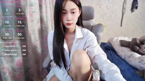 Media: A video of an Asian woman with long black hair, fair skin, and a slim physique, wearing a white button-down shirt, seated on a bed in a dimly lit room with floral curtains and cluttered surroundings.