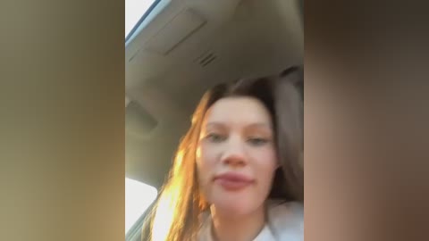 Media: Video of a woman with straight brown hair, fair skin, and a neutral expression, sitting in a car's passenger seat. The background shows car interior elements and sunlight streaming in.