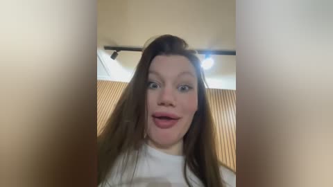 Media: Video of a young Caucasian woman with long brown hair, fair skin, and wide blue eyes, wearing a white top, in a room with beige walls and a wooden headboard.