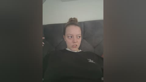 Media: Video of a young woman with light skin and brown hair in a bun, wearing a black sweatshirt, seated in a dark gray car interior, with a neutral expression.