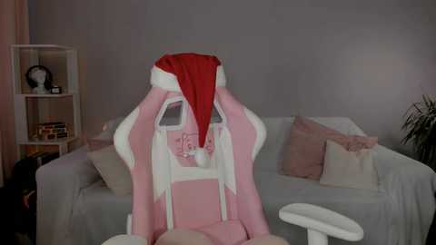 Media: Video of a pink gaming chair with a red Santa hat, set against a gray wall, with beige cushions and a shelf in the background.