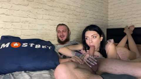 Media: Video of a bearded man with a beer belly, a tattooed woman giving him oral sex on a bed with a brick wall backdrop, blue pillow with \"Mostbet\" logo.