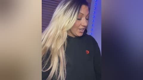 Media: Video of a blonde woman with long, straight hair, wearing a black top with a small red emblem, standing in a room with vertical blinds and blue LED strip lighting.