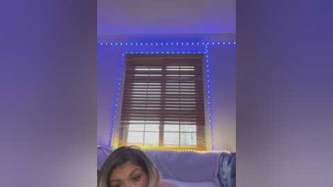 Media: Video of a woman with light skin, shoulder-length blonde hair, and a slim physique, seen from the waist up, sitting on a bed with white sheets. A window with wooden blinds and blue LED strip lights is behind her, creating a serene, intimate ambiance.