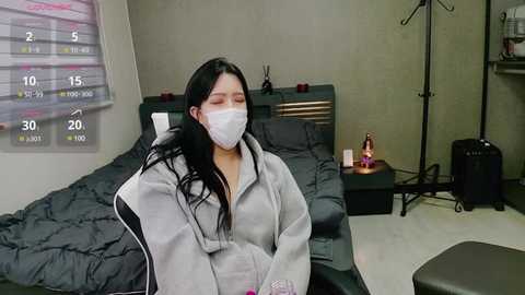 Media: Video of an East Asian woman with long black hair, wearing a white face mask and gray hoodie, sitting on a bed with gray sheets and a wooden headboard in a sparsely decorated room.