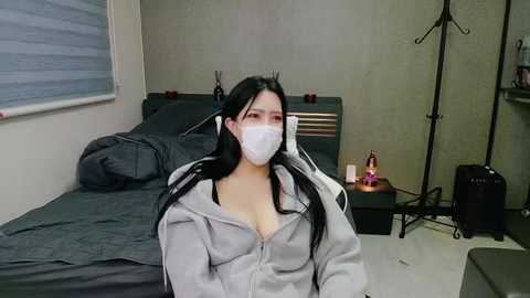 Media: Video of an Asian woman with long black hair, wearing a white mask and grey hoodie, seated on a bed in a dimly lit, minimalist bedroom with grey walls.