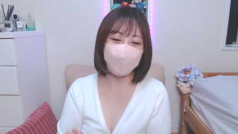 Media: Video of a young Asian woman with short dark hair, wearing a white mask and a white cardigan, seated in a pastel-colored room with a bed and dresser.