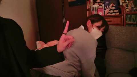 Media: Video of a woman with short dark hair, wearing a white mask and gray sweater, being forcefully pulled by the arm, sitting on a beige couch in a dimly lit living room with bookshelves in the background.