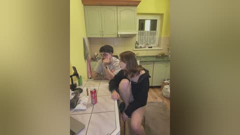 Media: A video shows a young man in glasses and a woman with long hair, both wearing casual clothes, sitting at a kitchen counter. The kitchen has light yellow walls and wooden cabinets.