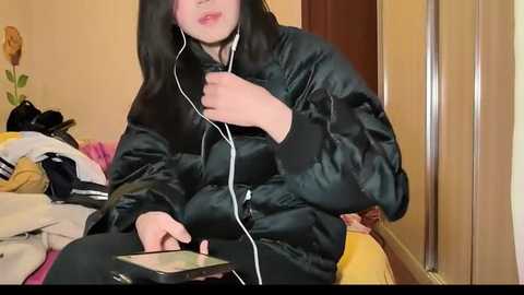 Media: Video of a person wearing a black puffer jacket, white earbuds, and listening to a smartphone. The background features a messy room with clothes on a bed and a vase of flowers.