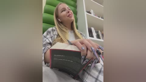 Media: Video of a young woman with long blonde hair, wearing an oversized plaid shirt, reading a book while seated on a green cushion. Background includes a white bookshelf filled with books and a green wall.