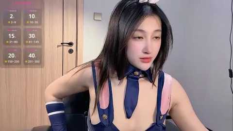 Media: A video of a young Asian woman with straight black hair, fair skin, wearing a revealing blue bunny costume with pink accents, seated indoors with a wooden door and calendar in the background.