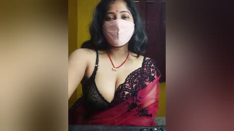 Media: A video of a South Asian woman with dark hair, wearing a pink mask, black lace bra, and a red saree with black floral embroidery, standing in a yellow room.