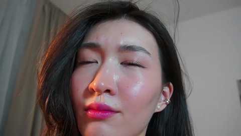 Media: Video of an East Asian woman with long black hair, closed eyes, and glossy skin, wearing earrings, indoors with beige walls and a curtain.