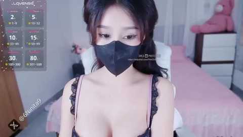 Media: A video of an East Asian woman with long black hair, wearing a black face mask, black lace lingerie, and a pink bra. She stands in a pink bedroom with a teddy bear, a bed, and a calendar.