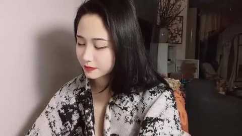 Media: Video of a young Asian woman with long black hair, wearing a black-and-white patterned robe, sitting in a dimly lit room with minimalistic decor and a white wall.