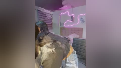 Media: Video of a person with long black hair, wearing a black jacket and jeans, standing in a dimly lit room with a pink neon sign on the wall and a partially visible bed.