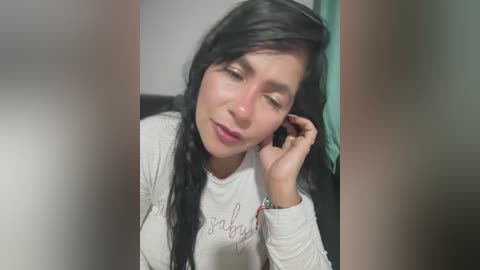 Media: Video of a Latina woman with long black hair, wearing a white long-sleeved shirt, sitting indoors, talking on a black phone, eyes closed, looking tired.