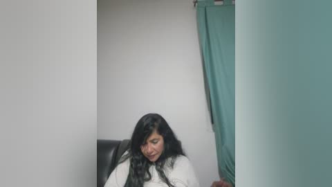 Media: Video of a woman with long black hair, wearing a white shirt, sitting in a dark office chair. Background includes white wall and teal curtain.