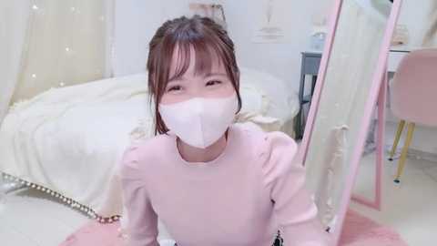 Media: Video of a young Asian girl in a pink dress, wearing a white face mask, sitting on a pink carpet in a softly lit, pastel-colored bedroom with white furniture.