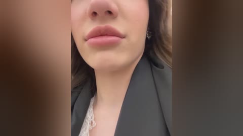 Media: Close-up video of a woman with light skin, straight brown hair, wearing a black blazer over a white lace top. Background is blurred, focusing on her face, lips, and neck.