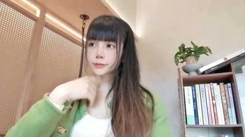 Media: Video of a young Asian woman with long, straight brown hair, wearing a green cardigan over a white top, standing in a modern, minimalist room with a bookshelf and potted plant in the background.