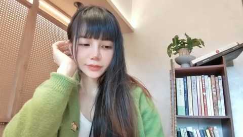 Media: Video of a young Asian woman with long black hair and bangs, wearing a green cardigan, standing indoors, adjusting her hair with one hand, while a bookshelf with books and a plant is visible in the background.