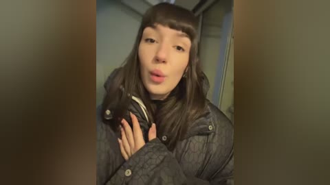 Media: A video of a young woman with long, straight brown hair and blunt bangs, wearing a puffy, dark grey jacket, blowing into a microphone. The background is a dimly lit, beige-colored room.