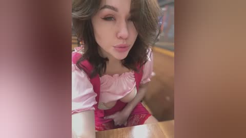 Media: A video of a young woman with wavy brown hair, wearing a pink dress, leaning over a wooden table in a dimly lit room.