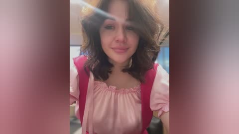 Media: Video of a young woman with wavy brown hair, wearing a pink blouse with ruffled neckline, sitting indoors. The image has a soft focus, with pink and brown gradient filters on the edges.