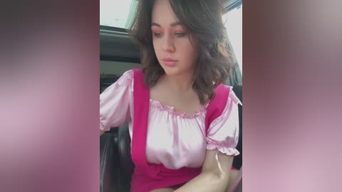 Media: Video of a young woman with wavy brown hair, wearing a pink top with ruffled sleeves, sitting in a car, looking downcast.