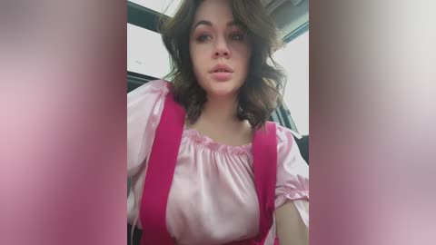 Media: Video of a young woman with shoulder-length wavy brown hair, wearing a pink blouse with puffed sleeves and a pink and white dress, seated in a car.