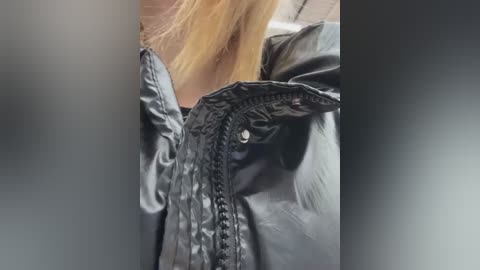 Media: Video of a woman's shoulder and hair, wearing a shiny, black, quilted leather jacket. Her long, straight blonde hair cascades down her back. The background is blurry, suggesting a focus on the jacket's texture and details.