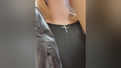 Media: A close-up video shows a woman's torso, wearing a black ribbed tank top with a silver cross necklace, partially obscured by a grey jacket. The background is blurred.