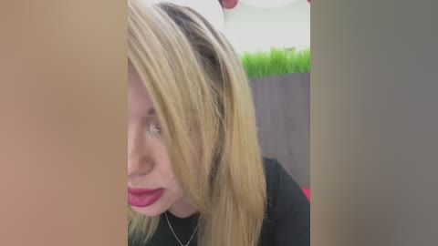 Media: Video of a blonde woman with long hair partially covering her face, wearing a black top, standing indoors with a green plant and beige wall background.